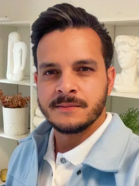 Ronney Ferreira de jaqueta azul-clara, no estúdio, com estante e esculturas ao fundo